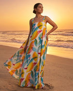 A woman stands on a sandy beach at sunset wearing a sleeveless maxi dress with a colorful tropical leaf print in yellow, orange, blue, green, and white.