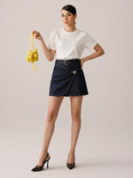 A woman modeling a white short-sleeve crewneck t-shirt and a dark navy blue wrap-style mini skirt with a silver buckle belt. She is holding a yellow mesh bag of lemons and wearing black pointed-toe stiletto heels, set against a solid light beige studio background.
