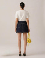 A woman is shown from behind wearing a white t-shirt and a dark blue denim mini skirt, holding a yellow mesh bag of lemons against a plain light beige studio background.