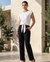 A woman modeling a white sleeveless top with a front tie knot and black wide-leg cropped pants, standing in a sunlit architectural setting with potted plants.