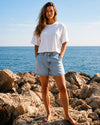 A woman stands barefoot on rocks by the sea, wearing a loose white short-sleeve T-shirt and light-wash denim shorts.